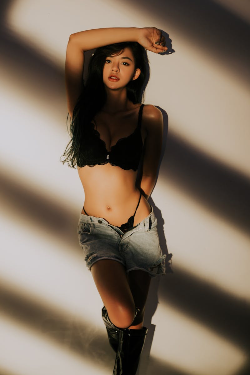 Sensual young slim female in elegant lingerie and denim shorts leaning on wall with arm above head and looking at camera in sunlight